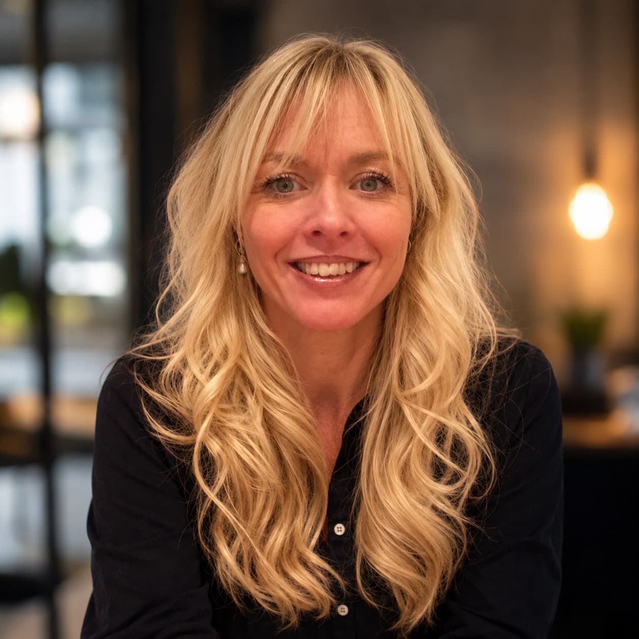 Susanne smiler, med langt, lyst og bølget hår, pandehår og sort skjorte